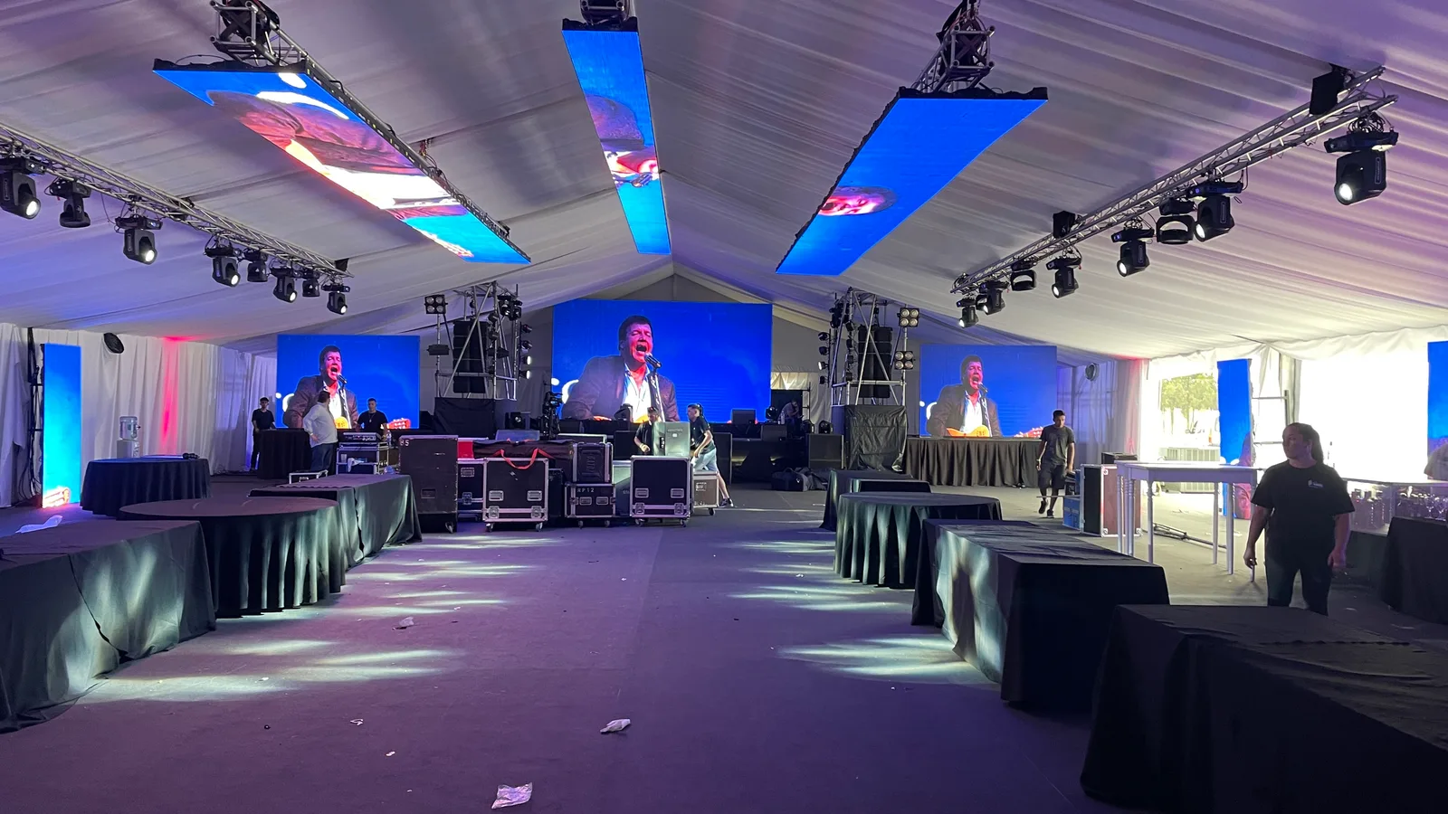 Escenario al fondo de una carpa con pantallas y mesas a ambos lados.