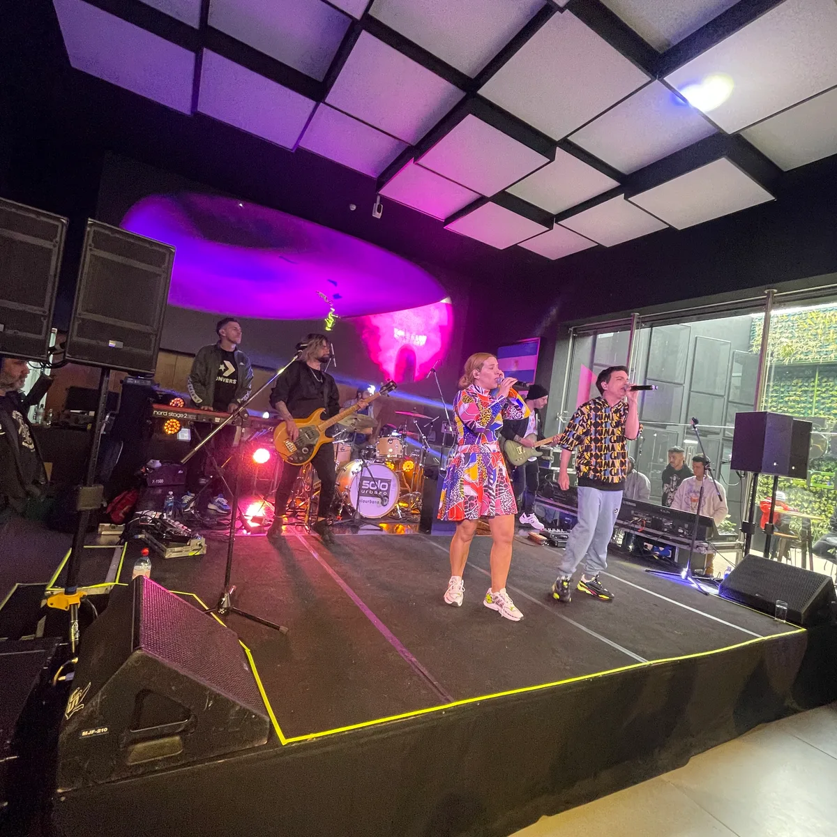 Banda musical sobre una tarima en un salón con ventanales.